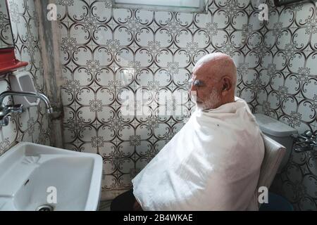 Very Old, White Haired, Bald Man waits for his Haircut In Old Vintage retro style tiled Bathroom Stock Photo