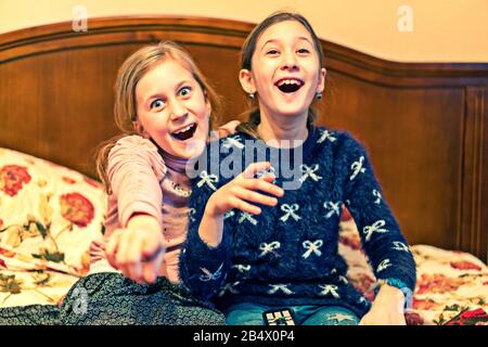 Two happy girls watching tv or movie. A horizontal photo of two young pretty smiling sisters lying on bed and watching their favourite serials. Stock Photo