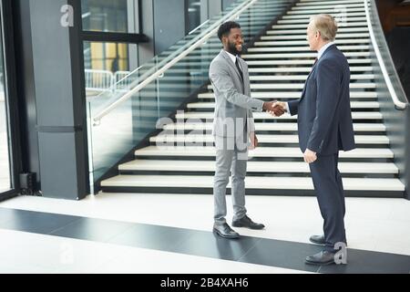 Horizontal full shot of two cheerful modern businessmen standing in front of each other shaking their hands Stock Photo