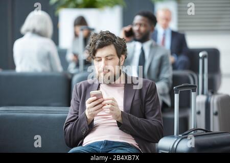 Horizontal portrait of young Caucasian man sitting relaxed in departure lounge surfing Internet on his smartphone Stock Photo