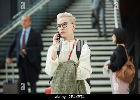 Horizontal portrait shot of stylish young woman standing in airport hall talking on phone, copy space Stock Photo