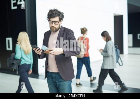 Young Caucasian man wearing eyeglasses standing in airport hall checking information about his flight on boarding pass Stock Photo