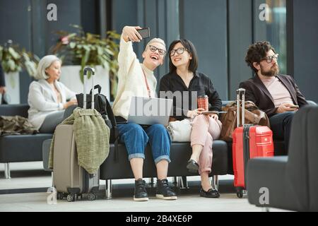 Two stylish young women sitting together in departure lounge taking selfie photo on smartphone camera, horizontal full shot, copy space Stock Photo
