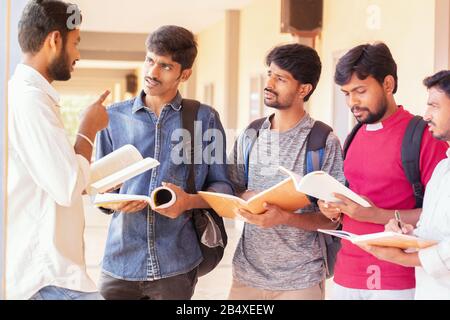 group of students listening to senior student to clear doubts - young intelligent man explaining to his classmates by looking into books at college - Stock Photo
