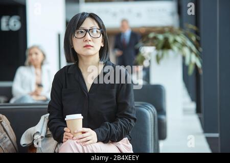 Sad young Asian woman wearing black shirt sitting alone in airport departure lounge holding cup of coffee looking away Stock Photo