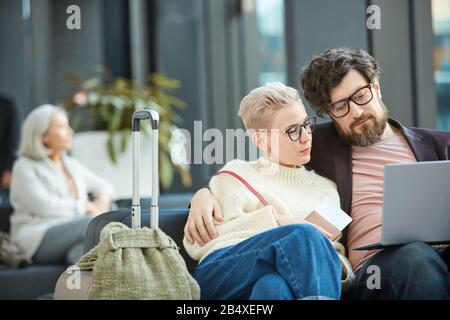 Young man and woman in love sitting together in departure lounge waiting for flight watching something on their laptop, copy space Stock Photo