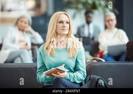 Horizontal medium portrait of mature blond woman sitting in depature lounge holding ticket and documents looking away Stock Photo