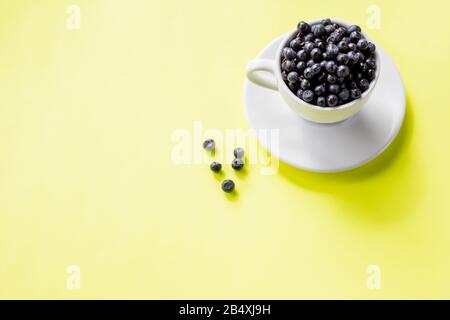 Ripe blueberries in a white coffee cup and a pattern of berries on a yellow background, copy space. Focus on blueberries in a cup.Fresh raw organic Stock Photo