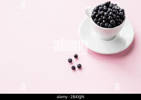 Ripe blueberries in a white coffee cup and a pattern of berries on pink background, copy space. Focus on blueberries in a cup.Fresh raw organic farm Stock Photo