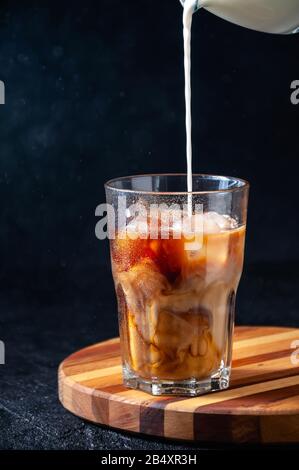 Milk Being Poured into Iced Coffee in Tall Glass on Dark Background. Concept Refreshing Summer Drink. Stock Photo