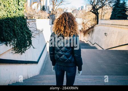 Rear view young woman with long curly hair stands at the top of staircase in city on sunny spring day. Stock Photo