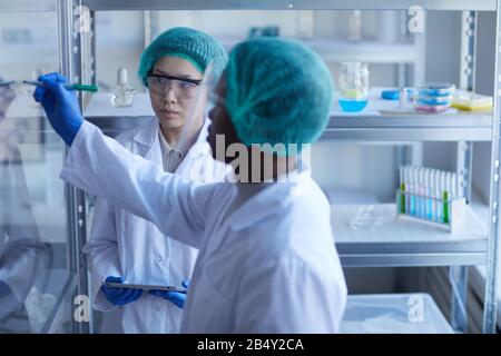 Horizontal portrait of Black male laboratory worker writing formula on glass wall, his female Asian colleague looking at it Stock Photo