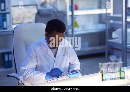 Horizontal portrait of African-American man wearing white coat and protective gloves sitting at desk in modern laboratory making notes Stock Photo