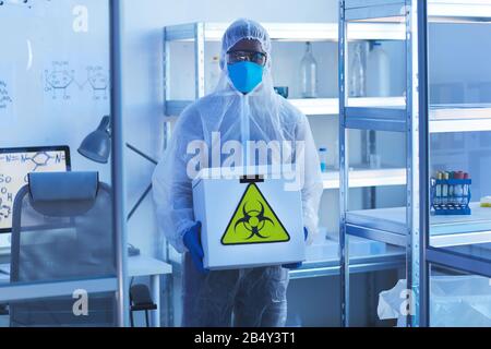 Horizontal medium long shot unrecognizable male laboratory worker in protective workwear holding box with biological hazards Stock Photo