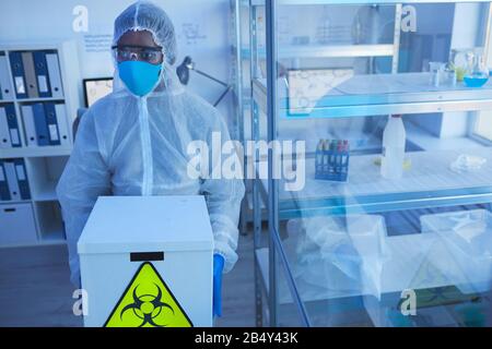 Horizontal high angle shot unrecognizable male laboratory worker in protective workwear carrying box with biological hazards Stock Photo