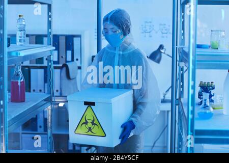 Horizontal medium long shot unrecognizable female laboratory worker in modern protective uniform holding box with biohazards Stock Photo