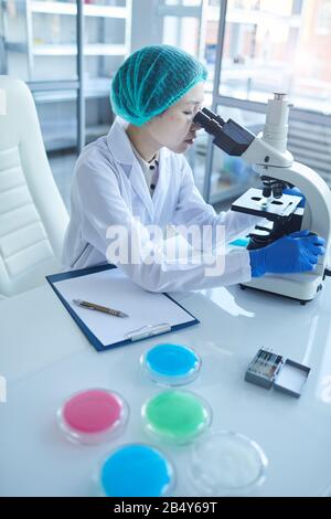 Vertical high angle portrait of young Asian female scientist analyzing liquid content using microscope, copy space Stock Photo