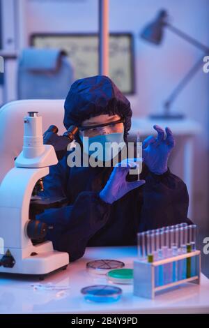 Vertical portrait of unrecognizable young laboratory worker in protective suit, eyewear and mask holding syringe with blue liquid medicament Stock Photo