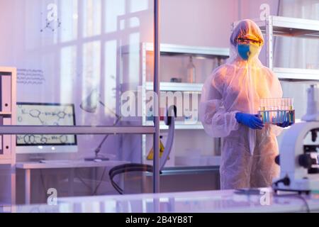 Unrecognizable female chemist wearing disposable protective uniform holding test-tubes with liquids looking at camera, horizontal medium long shot, co Stock Photo