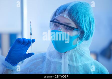 Horizontal head and shoulders close up shot of unrecognizable female medical laboratory worker wearing protective suit holding syringe with new vaccin Stock Photo
