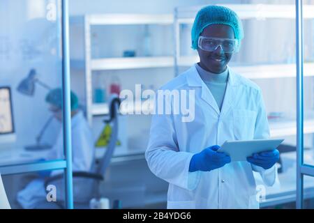 Horizontal medium portrait of cheerful young male scientist wearing white lab coat, medical cap and protective eyewear holding tablet PC looking at ca Stock Photo
