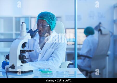 Horizontal shot of African American male medical scientist working in modern laboratory using microscope and making notes, copy space Stock Photo