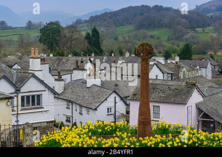 A war memorial surrounded by daffodils in the grounds of St Michaels and All Angels Church with the village of Hawkshead, Cumbria, in the background. Stock Photo