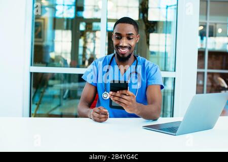 Portrait of a friendly male doctor or nurse wearing blue scrubs uniform and stethoscope sitting at desk with laptop in hospital checking mobile phone Stock Photo