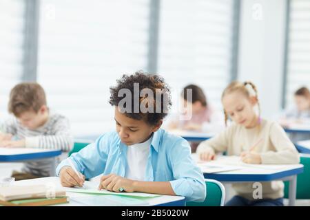 Horizontal medium close up shot of concentrated African American boy sitting at desk in classroom writing composition, copy space Stock Photo