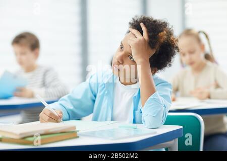 Pensive African-American boy wearing white T-shirt and blue shirt sitting at school desk doing difficult task, horizontal shot Stock Photo