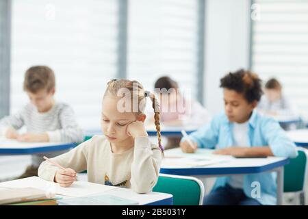 Horizontal portrait of little blond girl with two plaits sitting at school desk resting her cheek on hand learning boring lesson, copy space Stock Photo