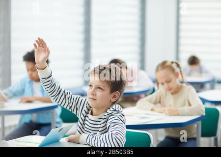 Ten-year-old boy wearing striped sweatshirt ready to give answer raising his hand in class, horizontal portrait, copy space Stock Photo