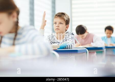 Horizontal portrai of modern primary school male student sitting at desk raising his hand, copy space Stock Photo