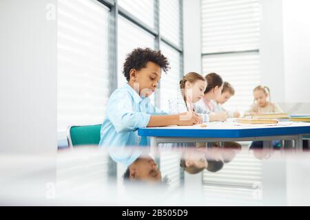 Pensive children wearing casual outfits sitting at school desk in modern classroom completing task in their notebooks, copy space Stock Photo