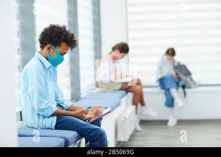 Horizontal side view shot of unrecognizable African American boy wearing mask playing mobile games while sitting in queque to school medical room, cop Stock Photo