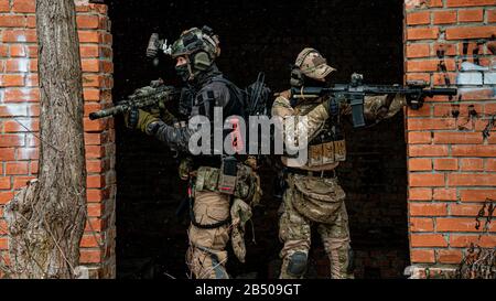 Two men in camouflage cloth and black uniform with machineguns stand back to back. Soldiers with muchinegun aims aiming from around the corner Stock Photo