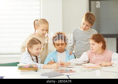 Two girls and boy sitting together in classroom drawing something in one notebook, their frends standing behind them, horizontal shot Stock Photo