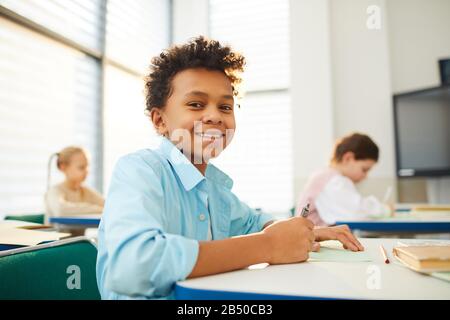 Horizontal low angle medium close up portrait of happy mixed-race boy with kinky hair sitting at school desk looking at camera smiling, copy space Stock Photo