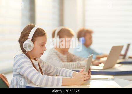 Middle school students sitting in classroom using modern headphones and computers during lesson, horizontal shot, copy space Stock Photo