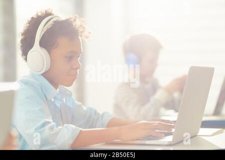 Medium side view portrait of modern African American boy wearing headphones using laptop during lesson time, copy space Stock Photo