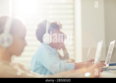 Medium side view portrait of modern twelve-year-old Black boy wearing headphones using laptop during lesson time, copy space Stock Photo