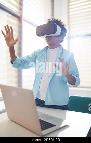 Vertical portrait of unrecognizable twelve-year-old Black boy standing at table with laptop on it using VR headset, copy space Stock Photo