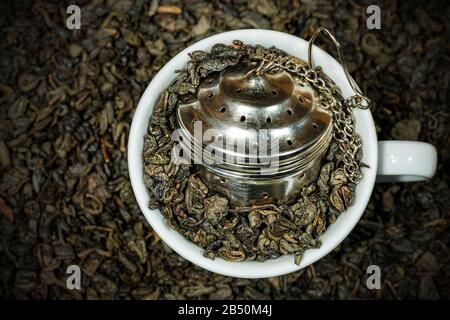 Close-up of dried leaves of green gunpowder tea in a white cup with a steel strainer. Many rolled up leaves on the background Stock Photo