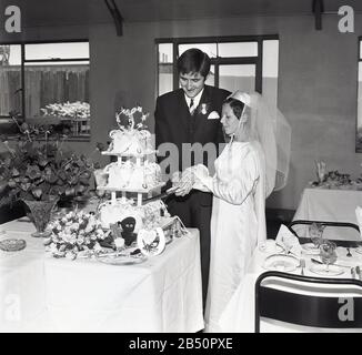 1960s, historical, a newly married couple holding hands as they stand cutting their wedding cake, England, UK. Stock Photo