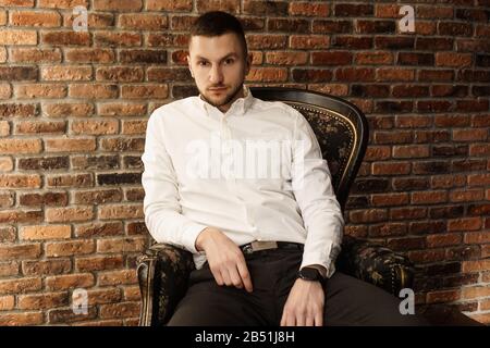 Portrait handsome fashionable man in a white shirt sits on a chair in a photo studio loft. Young businessman is thinking about future Stock Photo