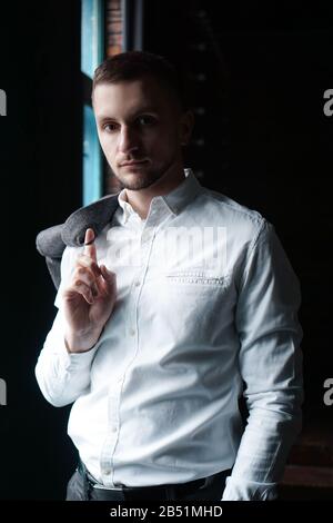 View in profile of a young businessman, dressed in a white shirt standing near the window on a dark walls and looking at the camera. Vertical photo Stock Photo