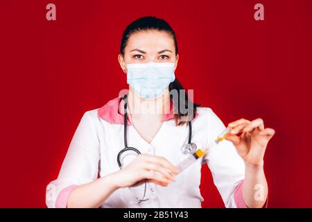 young girl doctor with a syringe in hand in a white coat and a mask on her face, fills a syringe on red background Stock Photo