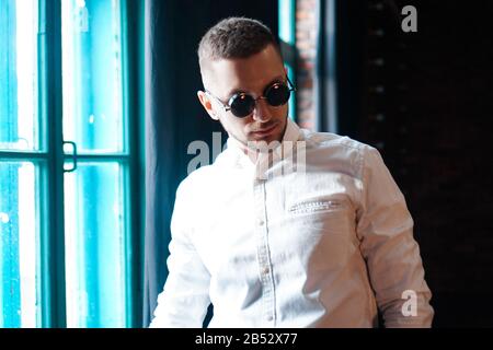 Handsome young man wearing vintage round black glasses looking forward on black background and window Stock Photo