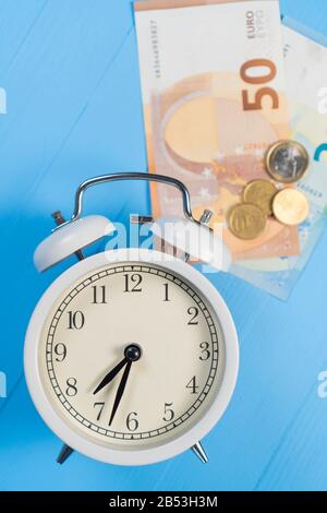 Alarm clock with euro banknotes and coins on a blue wooden background. Vertical shot. The concept of taxation and mortgage Stock Photo