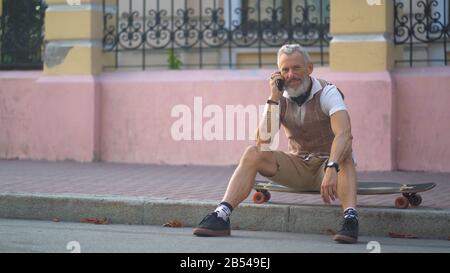 Middle-aged man communicates on the phone sitting on a skateboard in the city Stock Photo